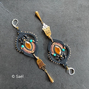 Könnte beinhalten: Ein Paar lange Makramee-Ohrringe mit schwarzer Kordel, bernsteinfarbenen ovalen Steinen und kleinen türkisfarbenen Perlen. Jeder Ohrring enthält einen goldenen Hamsa-Hand-Anhänger und einen tropfenförmigen Anhänger mit silbernen Klappbügeln.