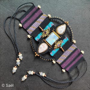 Puede incluir: Una pulsera de macramé con cordón negro, patrones tejidos en morado y turquesa, y una piedra rectangular iridiscente central flanqueada por dos piedras blancas en forma de lágrima. Incluye detalles de metal dorado y pequeñas cuentas blancas.