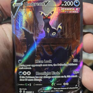 Puede incluir: Carta Pokémon Umbreon V holográfica. Presenta un personaje Umbreon negro con detalles amarillos, sobre un fondo oscuro que evoca una ciudad. El texto incluye "Umbreon V", "Single Strike" y descripciones de ataque.