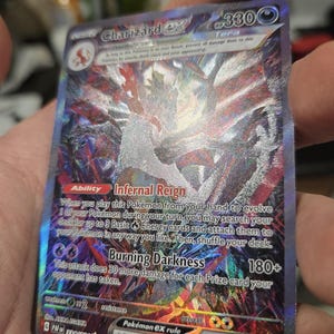 Puede incluir: Una carta coleccionable hologr&aacute;fica Charizard ex con 330 HP, que presenta el tipo Tera, la habilidad Infernal Reign y el ataque Burning Darkness. La carta muestra intrincados patrones de l&aacute;mina multicolor, ilustraciones oscuras y texto claro.
