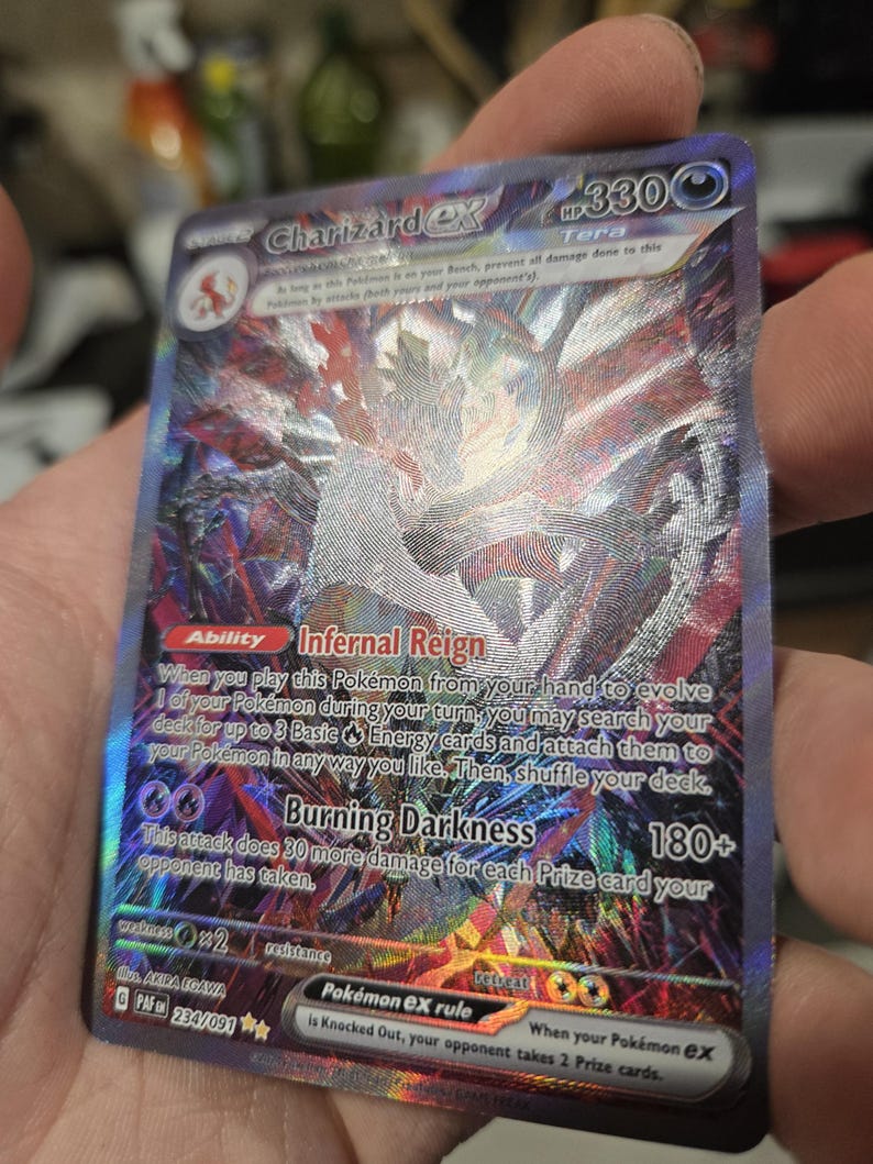 Puede incluir: Una carta coleccionable hologr&aacute;fica Charizard ex con 330 HP, que presenta el tipo Tera, la habilidad Infernal Reign y el ataque Burning Darkness. La carta muestra intrincados patrones de l&aacute;mina multicolor, ilustraciones oscuras y texto claro.
