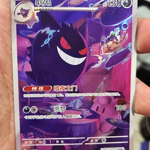 Op de afbeelding: Een holografische ruilkaart met een paars Gengar-personage met rode ogen en een brede grijns. De kaart heeft Japanse tekst en een HP-waarde van 130. De kaart wordt in een hand gehouden.