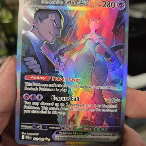 Puede incluir: Una carta holográfica de Pokémon con Mewtwo ex del Equipo Rocket. La carta muestra a Mewtwo morado y a un miembro del Equipo Rocket. El texto incluye las habilidades "Power Saver" y "Erasure Ball", junto con detalles de HP y ataque. La carta se sostiene en una mano.