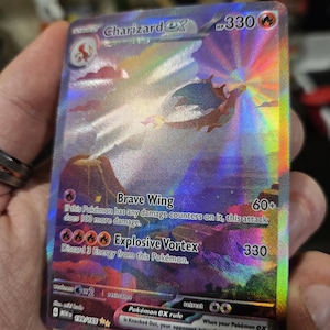 Puede incluir: Una carta coleccionable hologr&aacute;fica Charizard ex que muestra un paisaje vibrante con un Charizard volando, 330 HP y los ataques Brave Wing y Explosive Vortex. La carta muestra el texto Charizard ex, 199/165 y la regla de Pok&eacute;mon ex.
