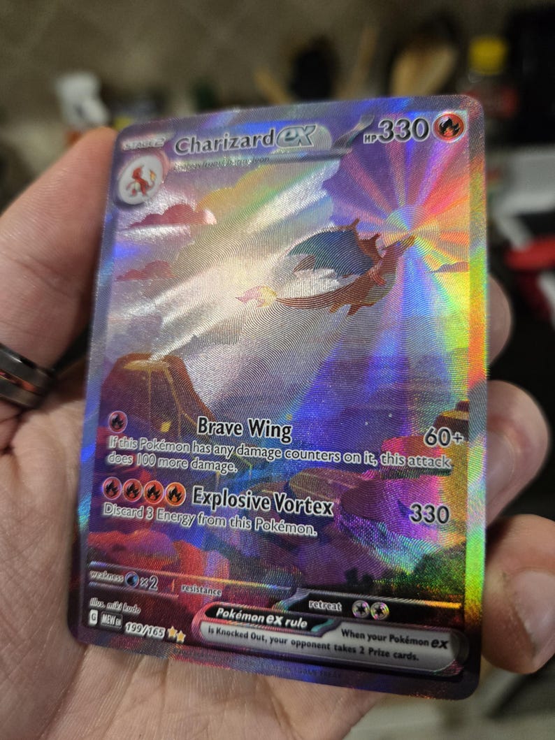 Puede incluir: Una carta coleccionable hologr&aacute;fica Charizard ex que muestra un paisaje vibrante con un Charizard volando, 330 HP y los ataques Brave Wing y Explosive Vortex. La carta muestra el texto Charizard ex, 199/165 y la regla de Pok&eacute;mon ex.