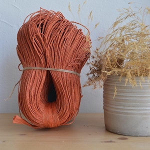 Può includere: Un primo piano di un rotolo di spago arancione, legato con uno spago marrone, su una superficie di legno. Uno vaso con erba secca è sullo sfondo.