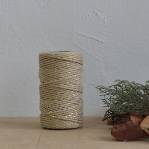 Peut inclure: Une bobine de ficelle naturelle avec des accents de paillettes dorées. La ficelle est enroulée autour d'un tube en carton et repose sur une surface brune.