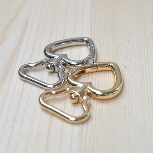 Könnte beinhalten: Drei herzförmige Metallschließen, zwei silberfarben und eine goldfarben, auf einer hellbraunen Holzoberfläche.