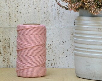 Könnte beinhalten: Eine Spule mit rosa Baumwollgarn liegt auf einer Holzoberfläche neben einem weißen Topf mit einer rosa blühenden Pflanze.