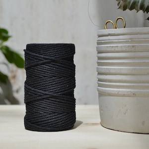 Może przedstawiać: Szpula czarnego sznurka bawełnianego, ciasno nawinięta, leżąca na drewnianej powierzchni. Sznurek jest gruby i fakturowany, a szpula ma około 12,5 cm wysokości.