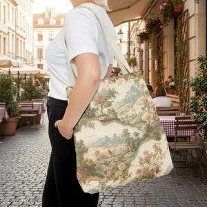 May include: A beige tote bag with a landscape toile pattern featuring trees, mountains, and a small house. The bag has a cream-colored strap and is carried over the shoulder. The bag is made of a sturdy fabric.