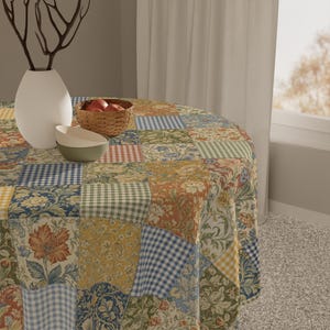 Puede incluir: Un mantel redondo de patchwork con una variedad de cuadrados de tela estampados florales, cuadros y otros, en tonos azules, verdes, amarillos y rojos. Un jarrón blanco, dos cuencos y una cesta de manzanas están sobre la mesa.