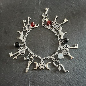 May include: A silver charm bracelet with a variety of charms, including keys, a snake, a crescent moon, a pentagram, and a witch's wand. The bracelet also features black and red beads.