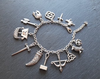 May include: A silver charm bracelet with various charms, including a sword, a hammer, a bow and arrow, a raven, a dragon, a ship, a knot, and a crown.