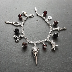 Op de afbeelding: Een zilveren armband met meerdere bedels, waaronder een raaf, een schedel, een zwaard, een spiraal en een veer. De armband heeft ook verschillende donkerrode glazen kralen.
