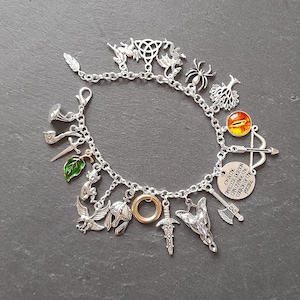 Puede incluir: Un brazalete de plata con varios dijes, incluyendo un dragón, un árbol, una araña, una espada, un anillo, un casco, un pájaro, una hoja, una pipa, un arco y una flecha, y un dije redondo con el texto "Incluso la persona más pequeña puede cambiar el curso del futuro".