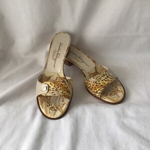 May include: A pair of beige heeled sandals with a leopard print and floral design. The sandals have a small buckle on the front strap and the brand name "Salvatore Ferragamo" is printed on the side.