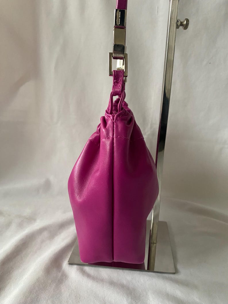 May include: A small, bright fuchsia-colored leather handbag with a silver chain strap. The bag is on a display stand.