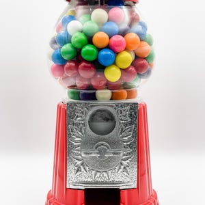 Puede incluir: Una m&aacute;quina de chicles roja con un globo de vidrio transparente lleno de chicles de colores. La m&aacute;quina tiene una ranura para monedas de plata y una palanca de plata.