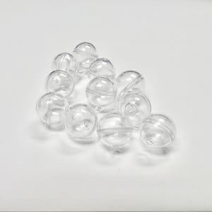 May include: A group of clear plastic beads, each with a small hole in the centre. The beads are arranged in a loose pile on a white surface.