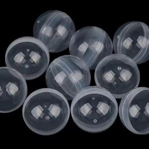 May include: A collection of clear, spherical plastic capsules, each split into two halves. The capsules are arranged against a black background, with light reflecting off their surfaces. These empty capsules are suitable for small toys or treats.
