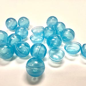 Puede incluir: Una colección de cápsulas y semiesferas de plástico azul translúcido. Las cápsulas son esféricas, con una costura alrededor del centro. Algunas tienen un diseño a rayas. Las semiesferas tienen forma de cuenco. Los objetos están dispersos sobre una superficie blanca.
