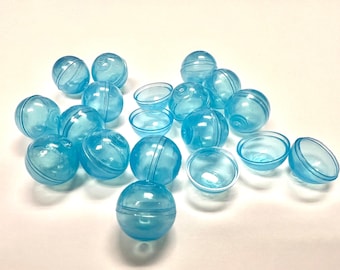 May include: A collection of translucent blue plastic capsules and half-spheres. The capsules are spherical, with a seam running around the middle. Some have a striped design. The half-spheres are bowl-shaped. The objects are scattered on a white surface.