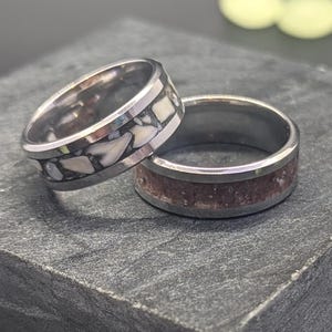 May include: Two silver rings with different inlays. One ring has a white inlay and the other has a brown inlay. The rings are on a gray surface.