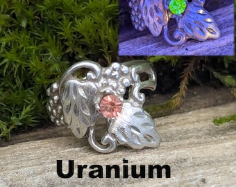 May include: A silver-toned ring with a grape and leaf design, featuring a pink gemstone. The ring is labeled "Uranium Glass". A smaller inset image shows the ring glowing green. The ring is on a wooden surface.