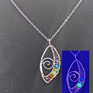 May include: A silver-toned wire-wrapped pendant necklace featuring a central spiral design and a vertical row of multi-colored faceted beads. The pendant is shown in standard lighting and also in a smaller inset image under UV light, which causes the beads to glow with a vibrant blue and purple luminescence.