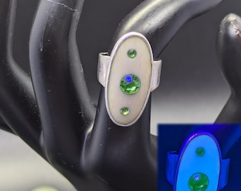 May include: Silver ring with an oval, off-white center stone. The stone is accented with three green and one blue gemstone. The ring is displayed on a black hand.