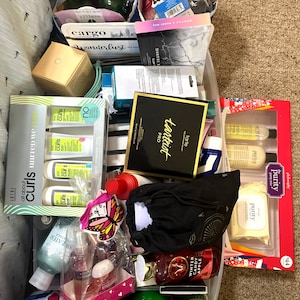 May include: A collection of various beauty and skincare products including boxed sets from brands like Philosophy, Tarte, and Curls. The assortment features bottles, tubes, palettes, and a black drawstring pouch, all packed together in a clear plastic bin.