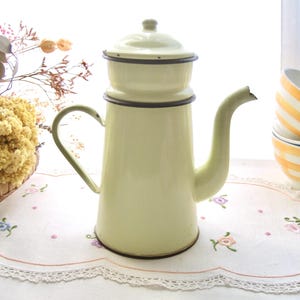 Könnte beinhalten: Eine cremefarbene Emaille-Kaffeekanne mit Deckel und gebogenem Ausguss. Die Kanne hat eine dunkelbraune Umrandung und einen passenden Griff. Sie steht auf einer weißen, mit Blumenmotiven bestickten Tischdecke. Im Hintergrund befindet sich ein Stapel gelb-weiß gestreifter Schalen.