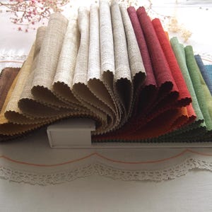 Puede incluir: Una variedad de muestras de tela en diferentes colores y texturas, incluyendo beige, marrón, rojo, verde y azul. Las muestras están dispuestas en forma de abanico y se muestran sobre una superficie blanca.