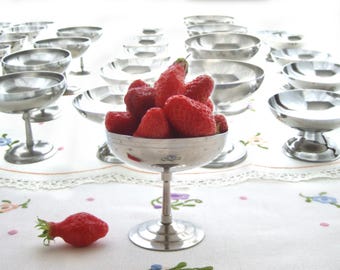 May include: A collection of silver-colored dessert dishes, some filled with fresh red strawberries. One dish is in the foreground, overflowing with berries. The dishes are arranged on a white tablecloth with floral embroidery.