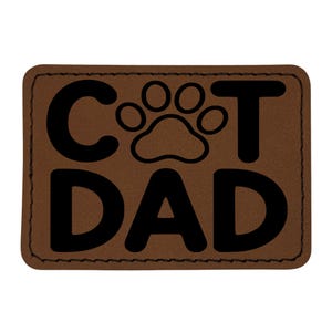 May include: A brown leather patch with the words "CAT DAD" in bold black letters. A black paw print graphic is between the words "CAT" and "DAD". The patch has a stitched border.