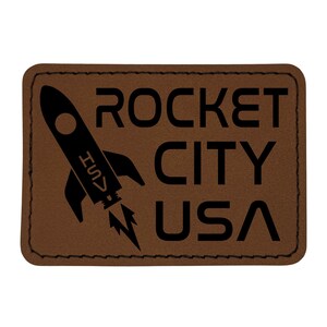 May include: A brown leather patch with the words "ROCKET CITY USA" in black block letters. A black rocket ship graphic is on the left side, with the letters "HSV" on the side of the rocket.