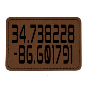 May include: A rectangular brown leather patch with black stitched edges. The patch features the coordinates 34.738228 and -86.601791 in bold, black font. The patch is likely a decorative item or a label.