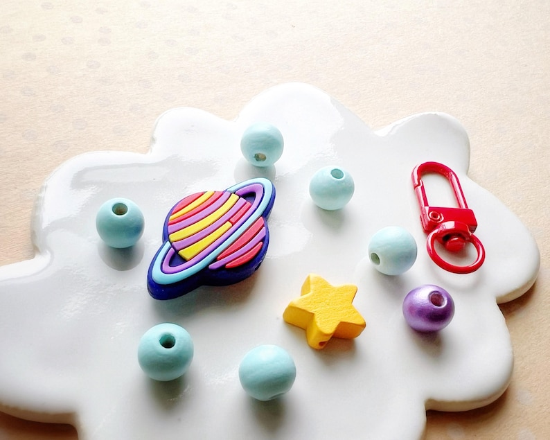 May include: A collection of colorful craft beads and charms. Includes light blue round beads, a planet-shaped charm with colorful stripes, a yellow star, a red clasp, and a purple bead. The items are arranged on a white, cloud-shaped surface.