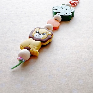 May include: A colorful silicone teether with a lion, a green leaf, and a red and orange bead. The teether is strung on a white cord.