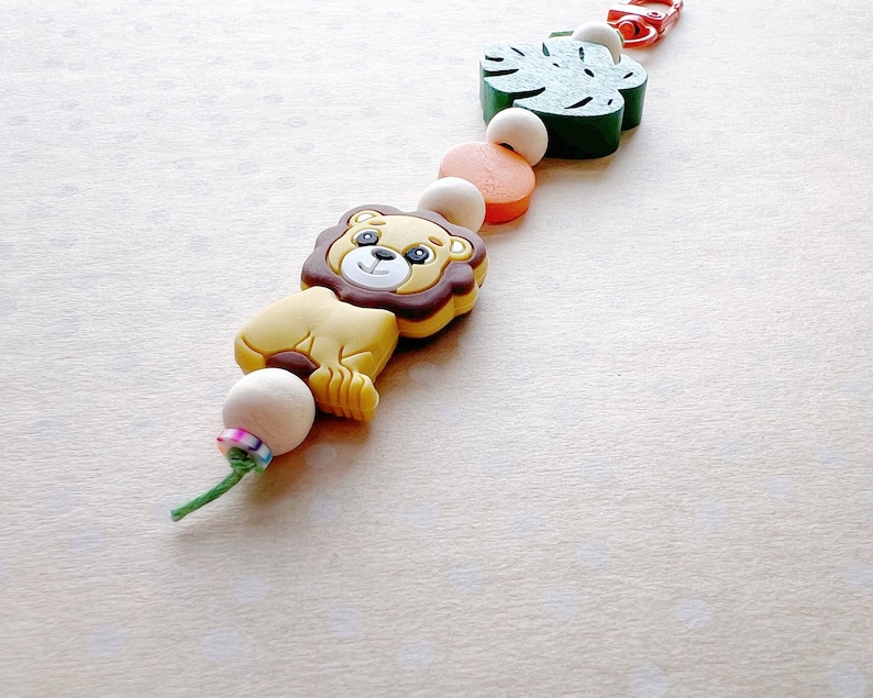 May include: A colorful silicone teether with a lion, a green leaf, and a red and orange bead. The teether is strung on a white cord.