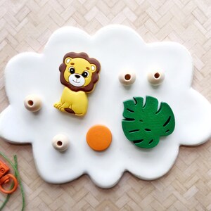 May include: A white cloud-shaped tray with a lion, a green leaf, wooden beads, and a small orange circle.