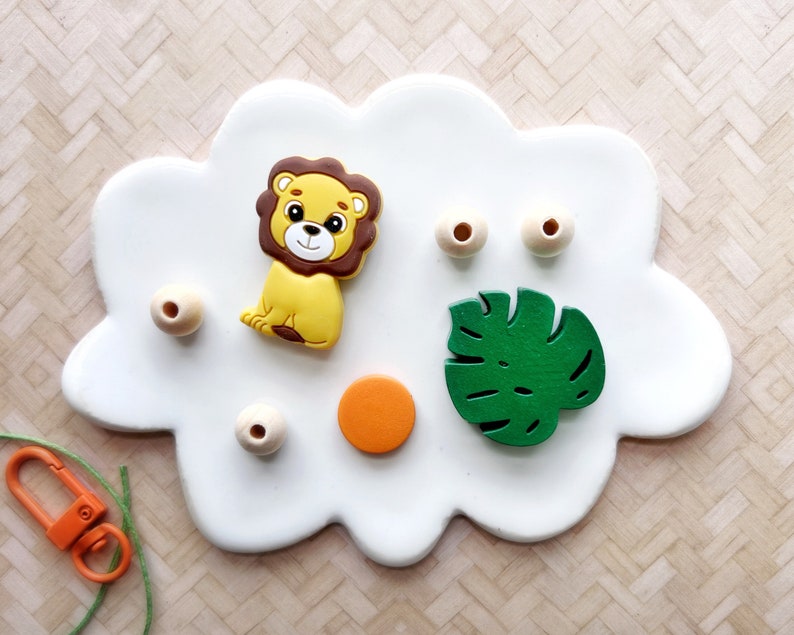 May include: A white cloud-shaped tray with a lion, a green leaf, wooden beads, and a small orange circle.