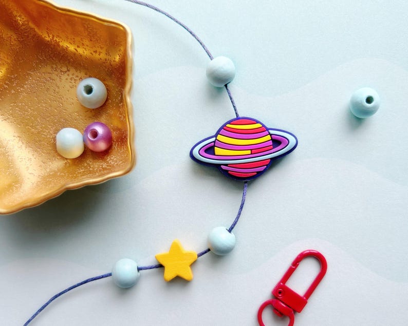 May include: A whimsical necklace featuring a planet charm with pink, yellow, and purple stripes, surrounded by a blue ring. The necklace includes a yellow star, blue beads, and a red clasp. The beads and planet charm are strung on a purple cord.