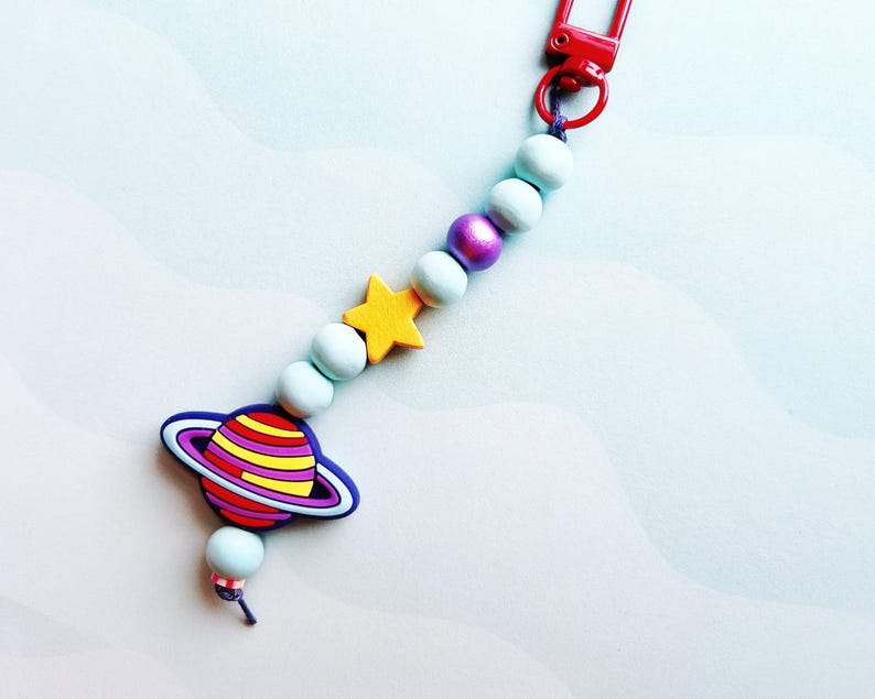 May include: A colorful keychain featuring a planet charm with a ring, a yellow star, and light blue beads. The keychain has a red clasp and a purple bead. The planet charm has a blue outline and a striped design in red, yellow, and purple.