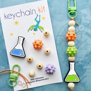 May include: A keychain kit with a white card that reads "keychain kit" with a cartoon illustration. The kit includes a green clasp, wooden beads, and colorful, spiky beads. A beaker-shaped charm with blue liquid is also included.