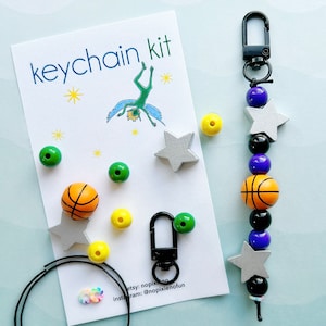 May include: A keychain kit featuring a white card with the text keychain kit and a green fairy illustration. The kit includes a black lobster clasp, black cord, and various beads including silver stars, orange basketballs, and purple, yellow, and green spheres.