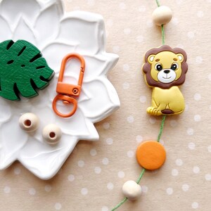 May include: A green leaf-shaped wooden cutout, two wooden beads, a bright orange key chain with a metal loop, and a yellow lion-shaped toy with brown mane on a green string with orange and wooden beads.