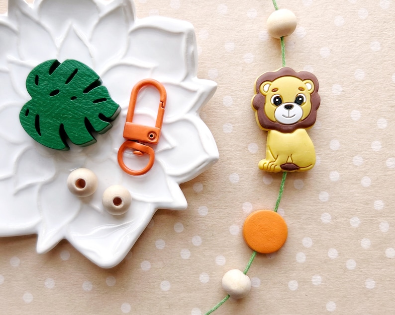 May include: A green leaf-shaped wooden cutout, two wooden beads, a bright orange key chain with a metal loop, and a yellow lion-shaped toy with brown mane on a green string with orange and wooden beads.