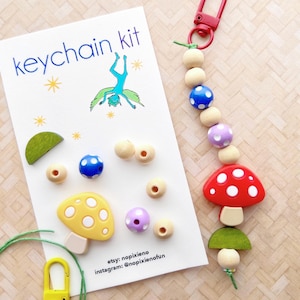 May include: A DIY keychain kit with wooden beads, mushroom charms, and a green string. The kit includes instructions for making a keychain with the text "etsy: nopixieno instagram: @nopixienofun".